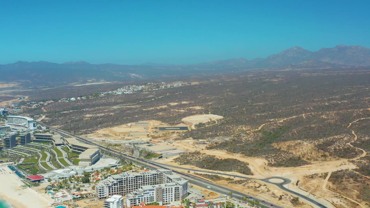 Mid-clip aerial view of Dreams Los Cabos with buildings in the lower foreground, a curved road and roundabout, sandy lots, hazy mountains, and no visible people.