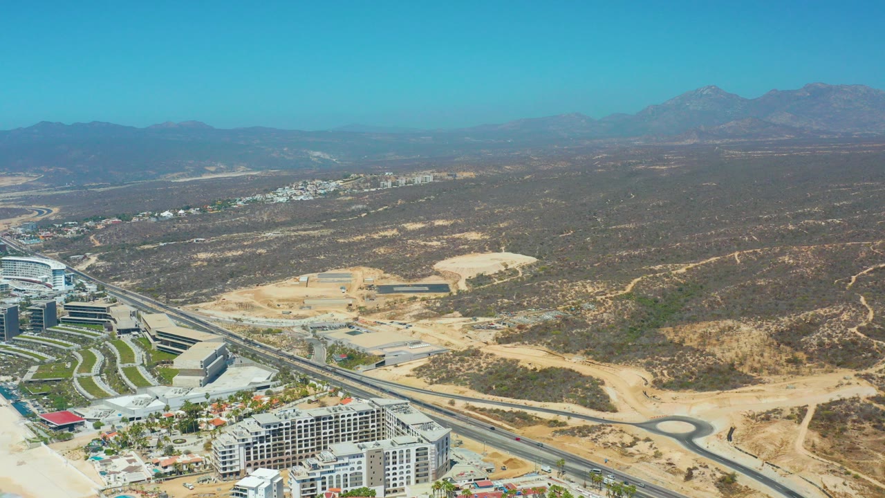 High aerial frame over Dreams Los Cabos showing highway lanes, resort-area buildings, tan construction ground, dry hills, distant mountains, clear blue sky, and no visible people.