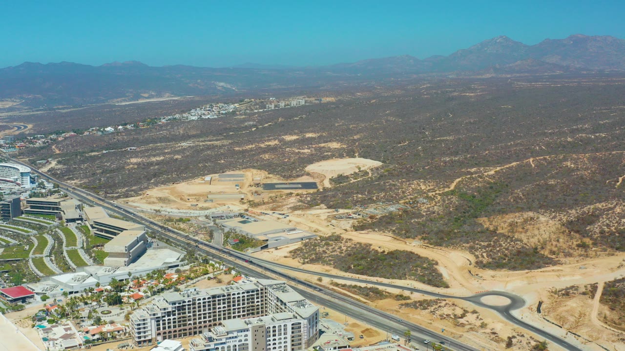 Opening aerial frame over Dreams Los Cabos with a highway and roundabout crossing sandy desert development, distant mountains under clear sky, and no visible people.