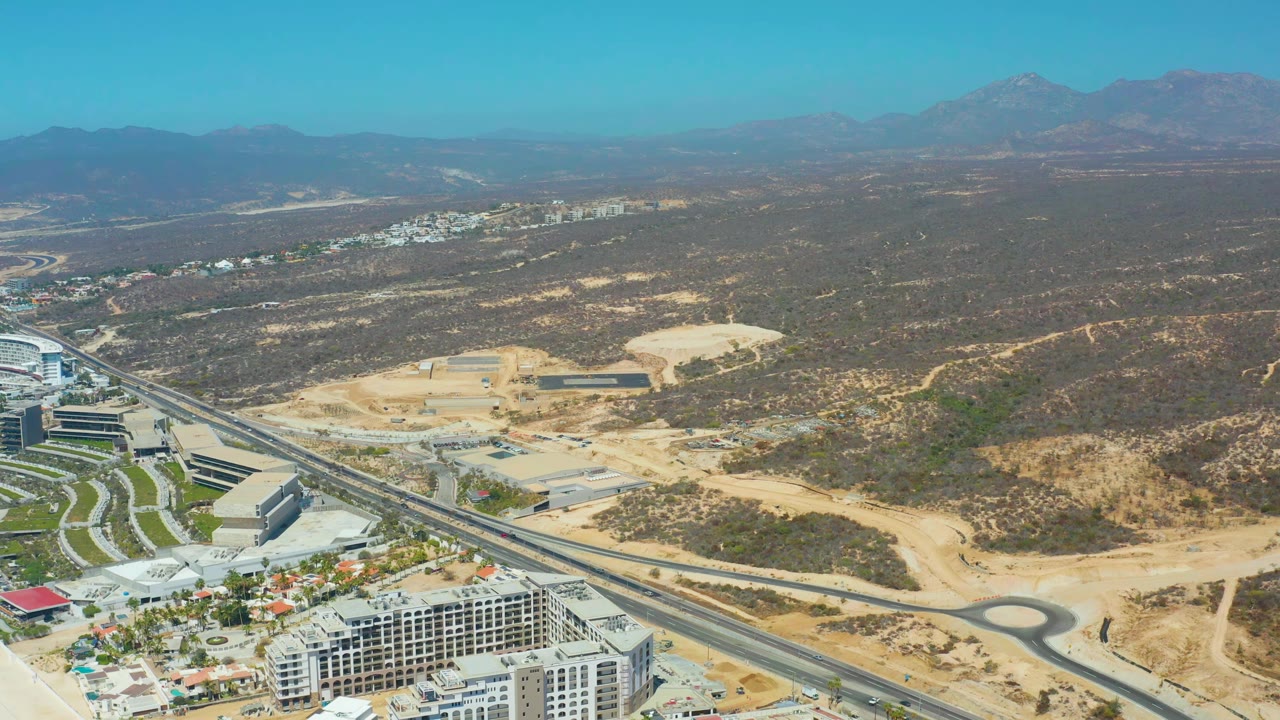 Aerial view in Dreams Los Cabos showing foreground buildings beside a divided highway, sandy graded land, desert vegetation, mountains, and no visible people.