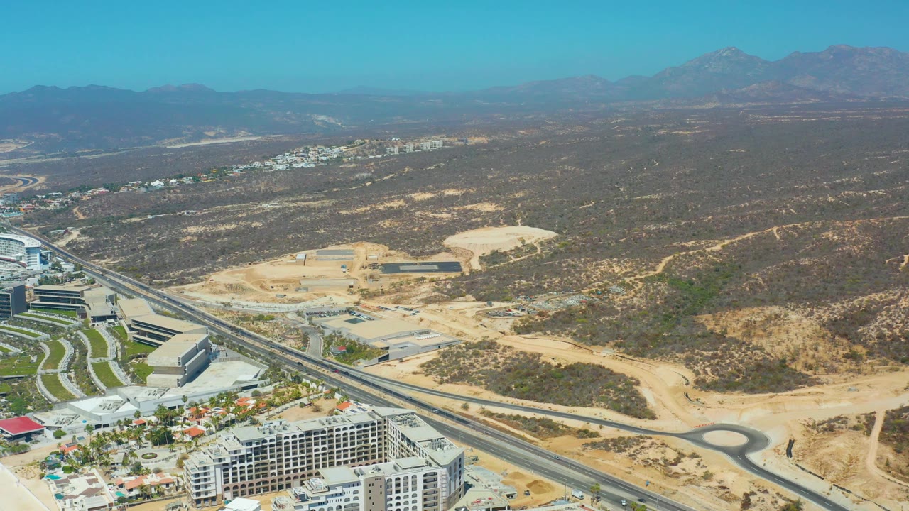 Drone-style frame over Dreams Los Cabos with the roundabout near the lower right, highway cutting across the scene, construction pads, scrubland, distant buildings, and no visible people.