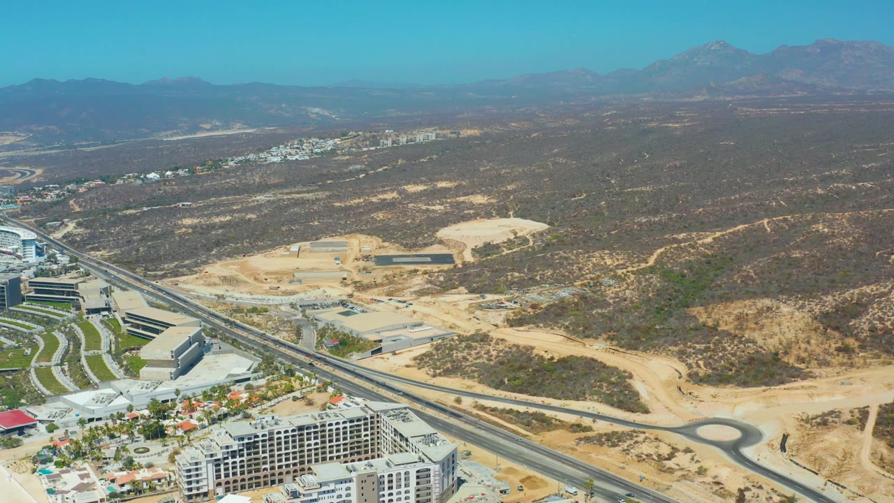 Wide aerial frame in Dreams Los Cabos with resort-area buildings at the lower edge, a highway and roundabout through sandy land, brown desert scrub, mountains, and no visible people.