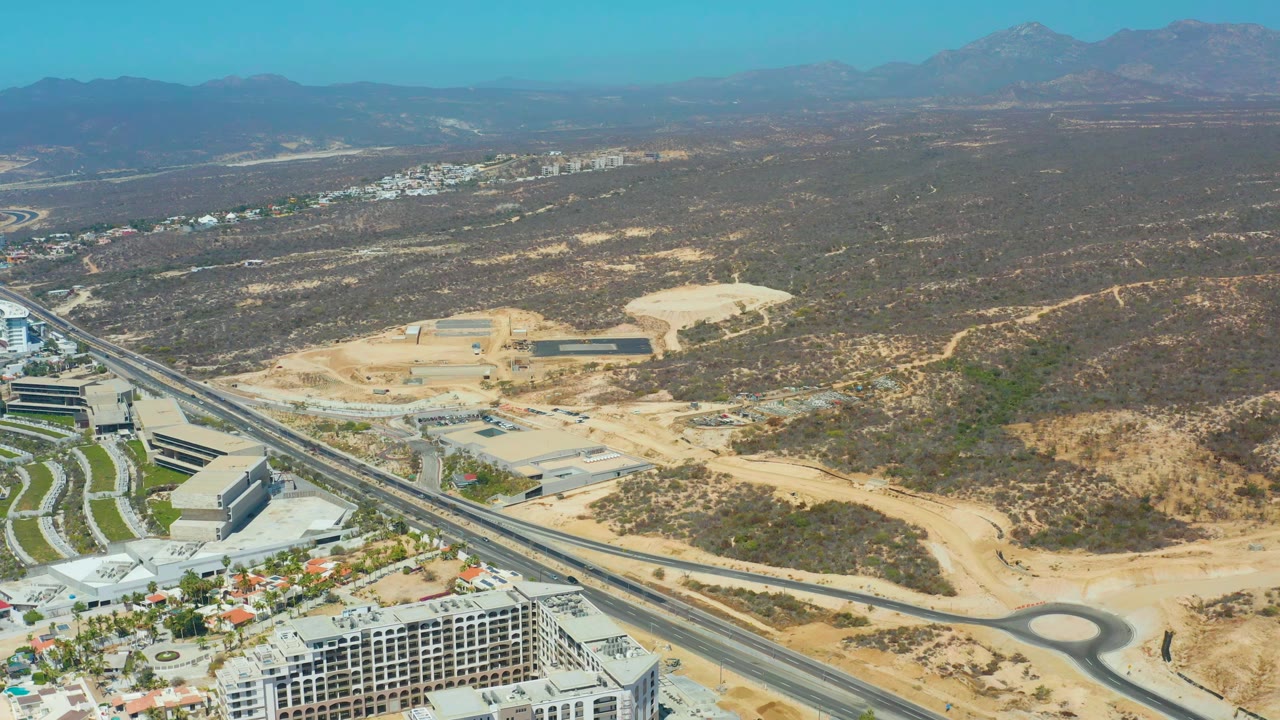 Drone-like aerial frame in Dreams Los Cabos showing the highway running diagonally past resort buildings, exposed construction ground, brown desert slopes, mountains, and no visible people.