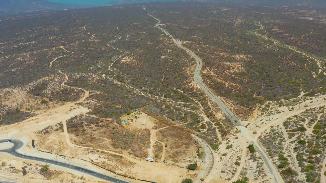 Aerial view near Dreams Los Cabos showing winding paved and dirt roads through arid hills, sparse vegetation, distant mountains, blue sky, and no visible people.
