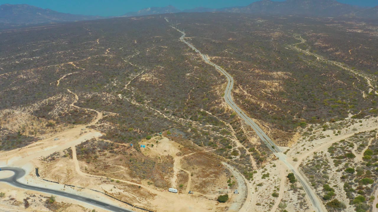 Aerial view near Dreams Los Cabos with the main road running from the lower frame into the hills, branching dirt tracks, dry scrubland, distant mountains, and no visible people.