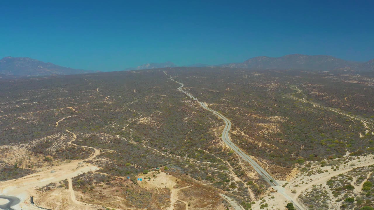Aerial view over Dreams Los Cabos terrain with the road angled through scrub-covered hills, tan dirt foreground, distant mountains, and no visible people.
