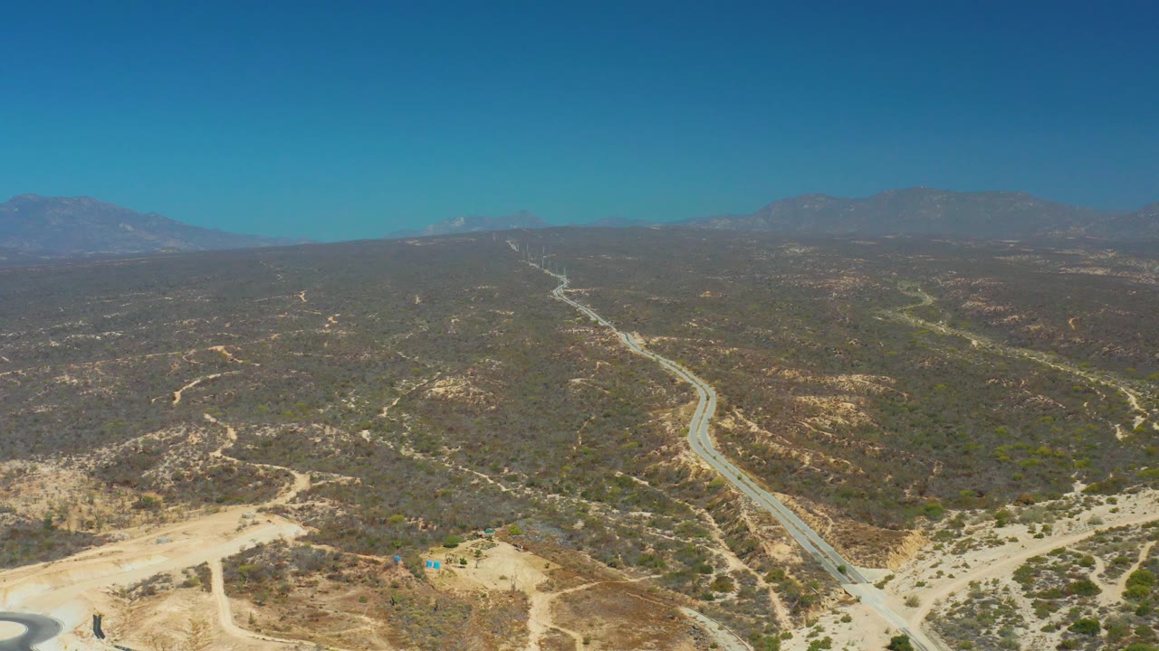 Aerial view from Dreams Los Cabos with a winding road through scrubby brown hills, pale dirt areas in the lower frame, mountain ridges beyond, and no visible people.