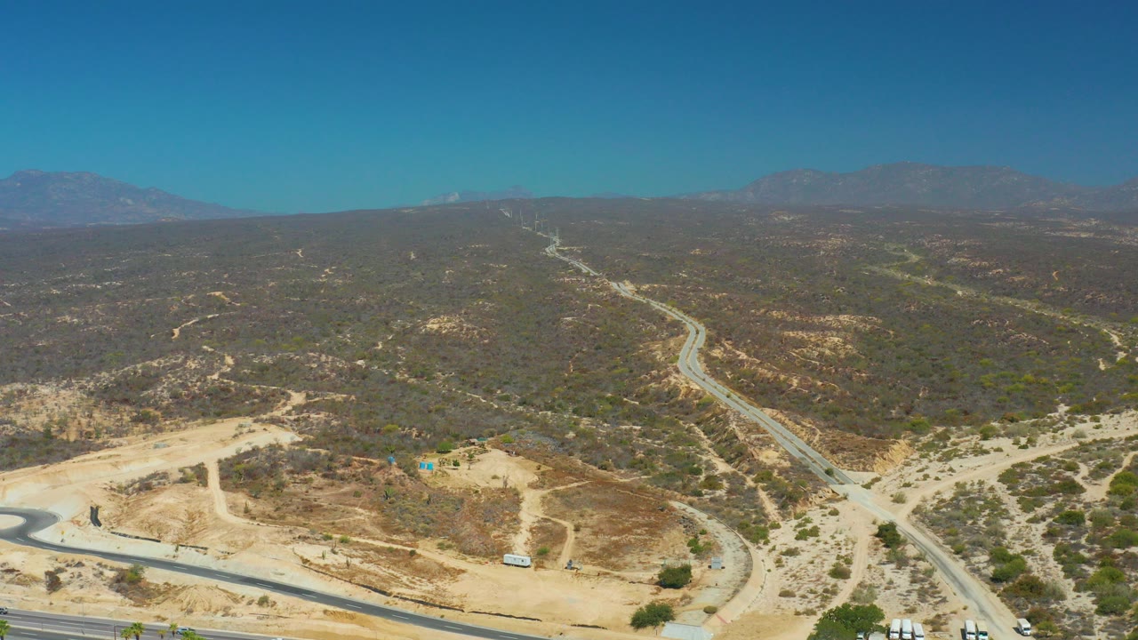 High aerial frame over Dreams Los Cabos showing a highway and winding road through dry hills under a clear blue sky, with no visible people.