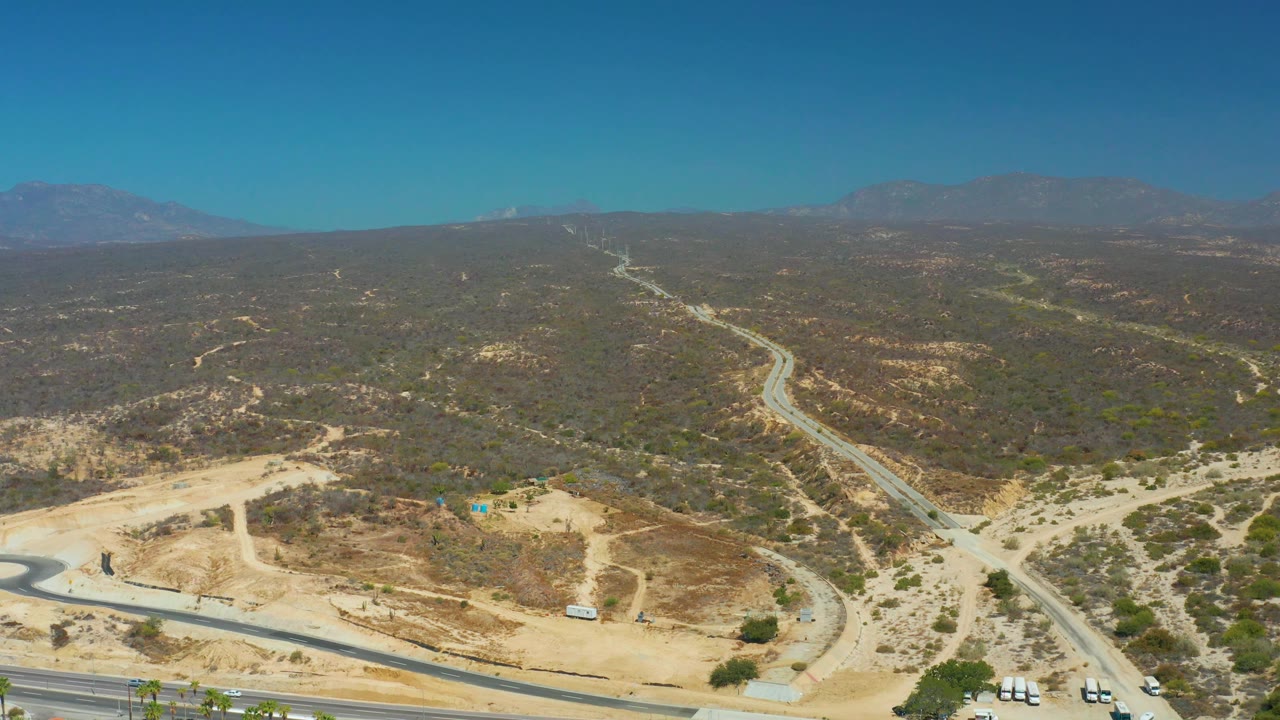 Aerial frame over Dreams Los Cabos showing a highway along the lower edge, a winding road rising into dry hills, distant mountains, and no visible people.