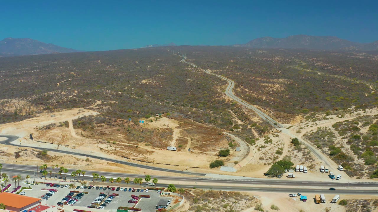 Aerial frame at Dreams Los Cabos with the highway crossing the lower image, sandy undeveloped ground and parked cars near the bottom, a winding road into scrub hills, distant mountains, and no visible people.