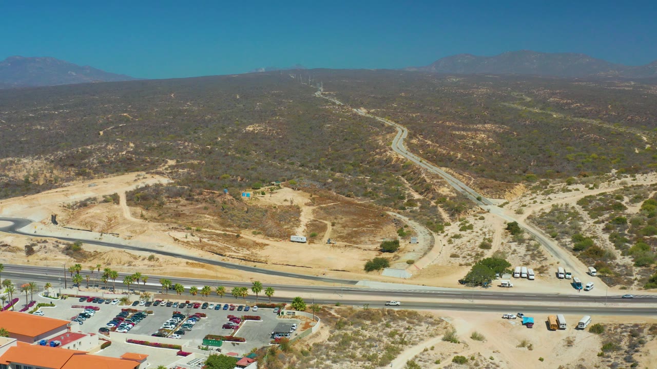 Mid-clip aerial view at Dreams Los Cabos with a red-roofed building corner and parking lot at the bottom, vehicles on the highway, sandy construction-like ground, scrubby hills, distant mountains, and no visible people.