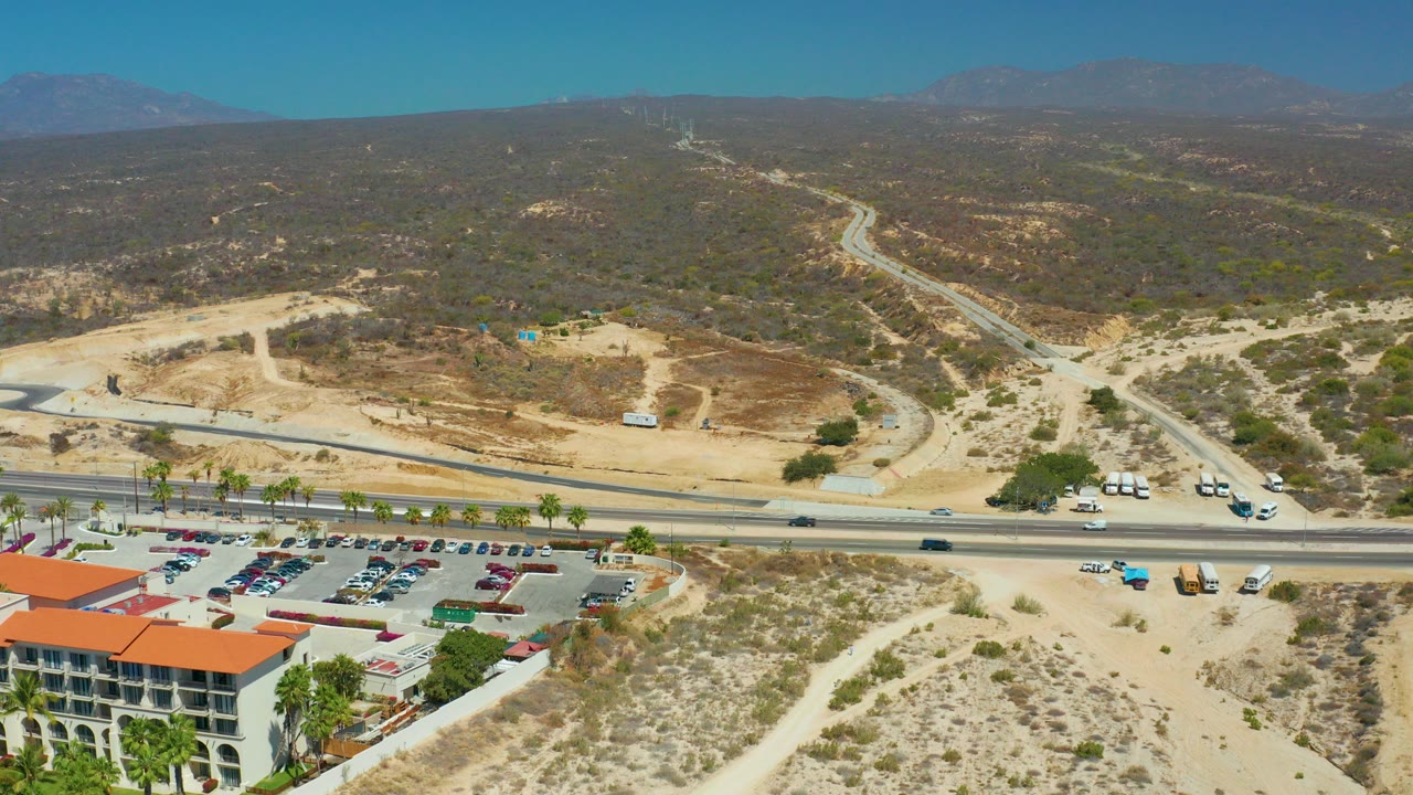 Opening aerial frame over Dreams Los Cabos shows a paved road, parked cars, a red-roofed building at the lower left, sandy scrub hills, distant mountains, and no visible people.