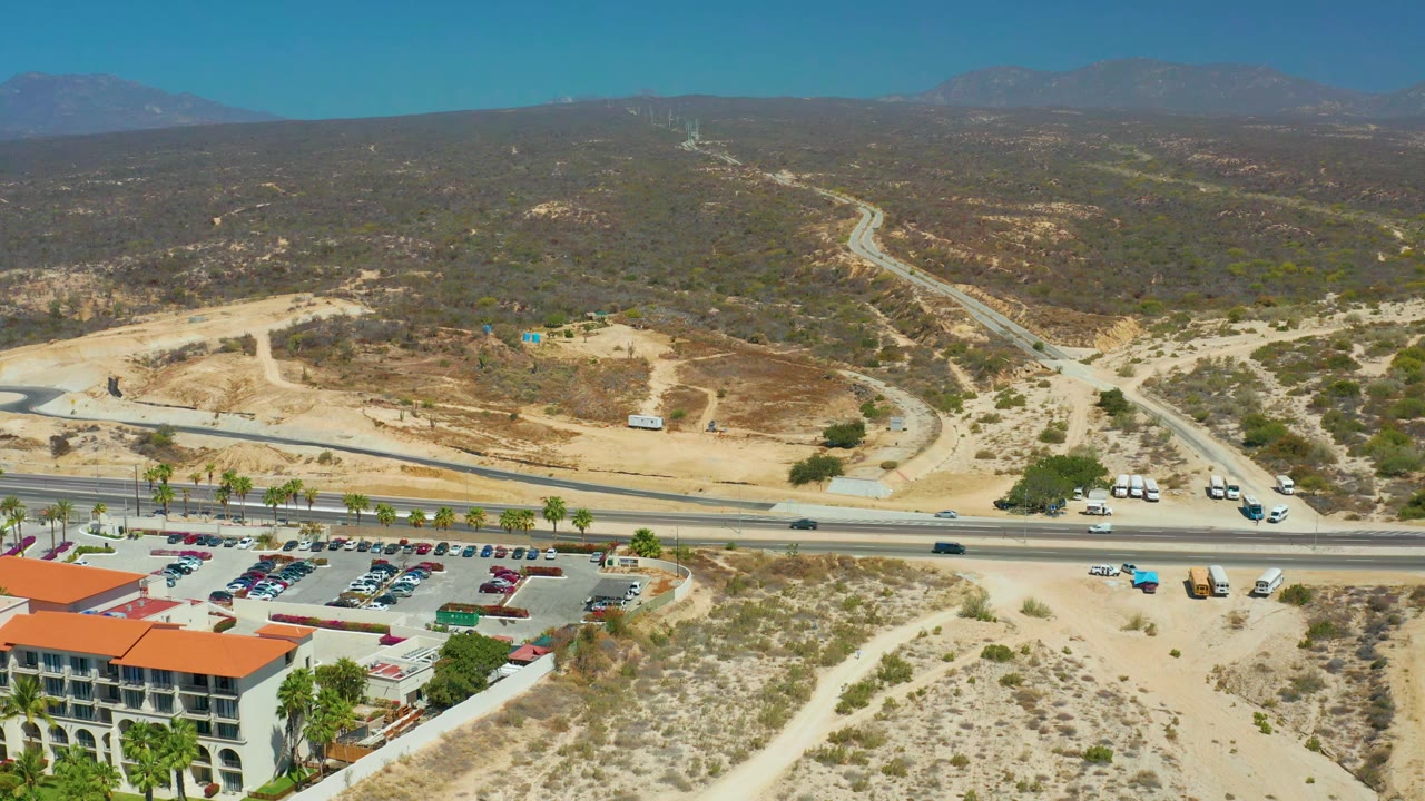 At 0:01, the aerial view over Dreams Los Cabos holds on the road and parking lot, with vehicles spaced along the pavement, pale sand hills beyond, and no visible people.