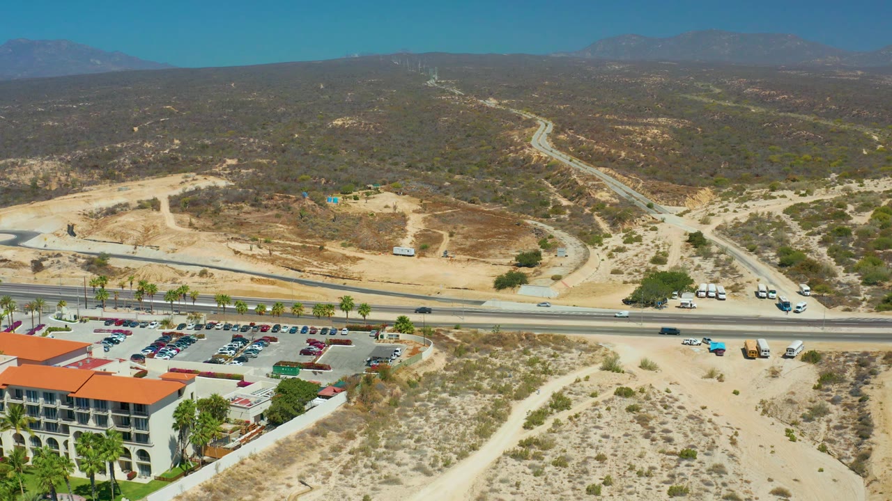 At 0:02, Dreams Los Cabos appears from above with a red-roofed building near the lower frame, a palm-lined parking lot, road traffic, sandy slopes, and no visible people.