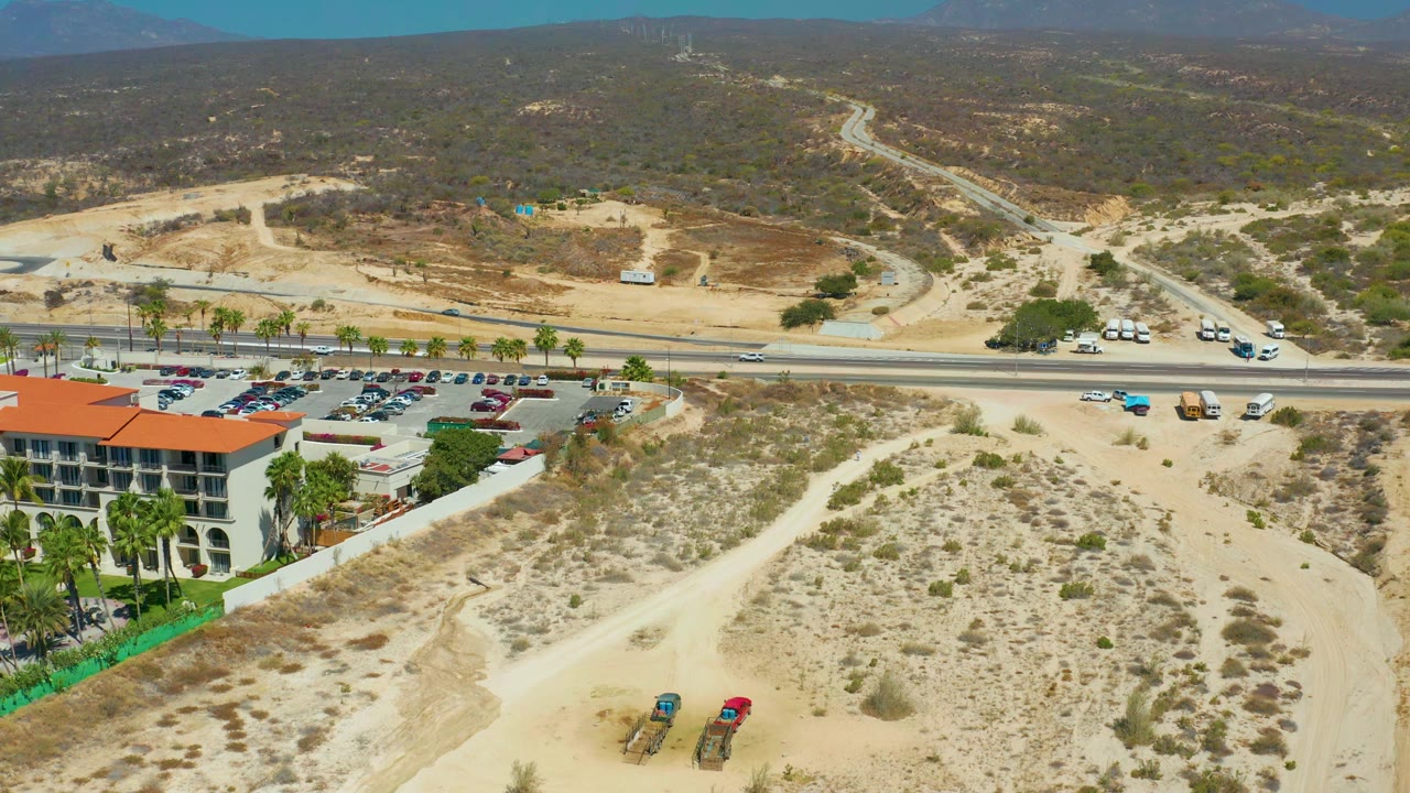Aerial view at Dreams Los Cabos showing dirt tracks and scrub in the foreground, two pieces of construction equipment below, a road and parking lot beyond, and no visible people.