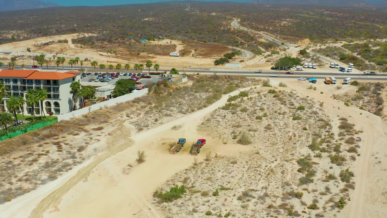 Opening aerial frame at Dreams Los Cabos showing sandy scrubland, a paved road with vehicles, a red-roofed building and palms on the left, two parked ATVs in the foreground, distant hills, and no visible people.