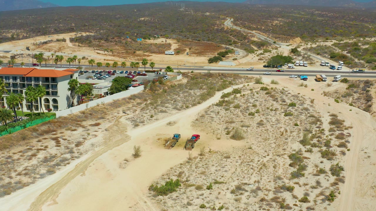 One second in, the Dreams Los Cabos view remains wide with light traffic on the road, the red-roofed building partly visible at left, parked ATVs on the sand, scrub patches, hazy hills, and no visible people.