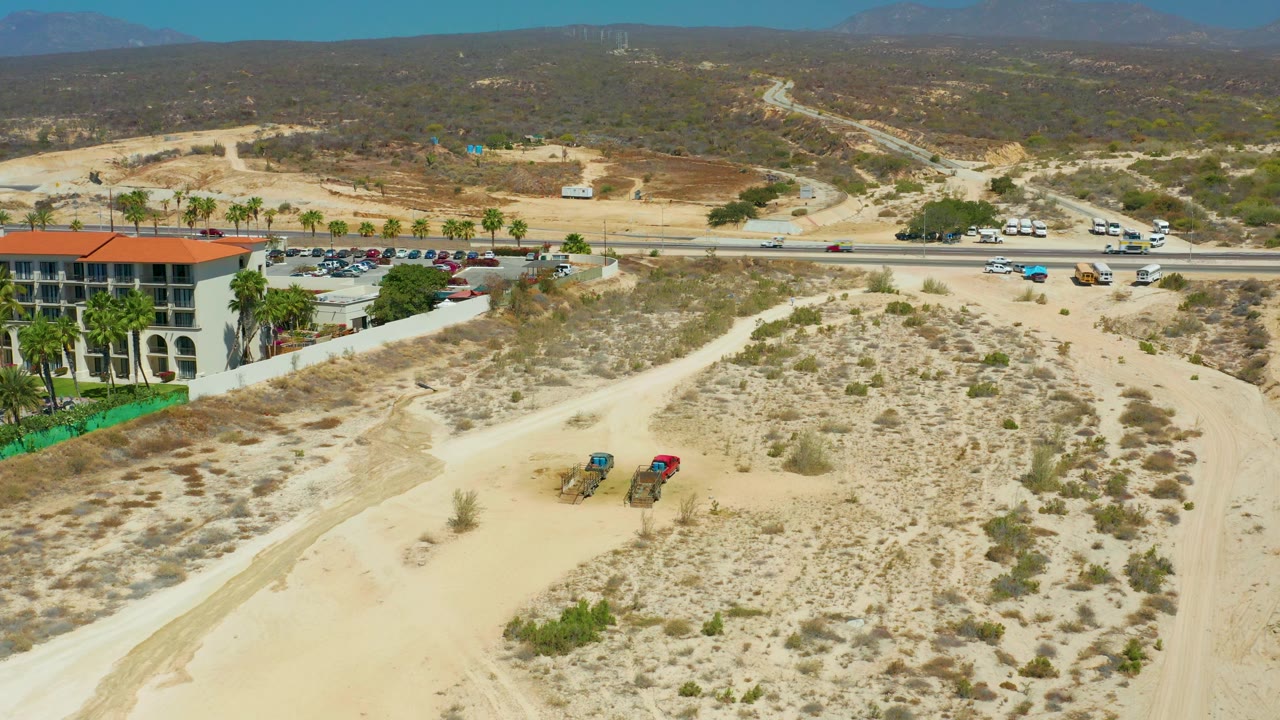 At 0:02, the aerial frame shows the sandy open lot and scrub leading to the road, with small vehicles traveling across the midground, a building and palms at left, distant hills, a bluer sky, and no visible people.