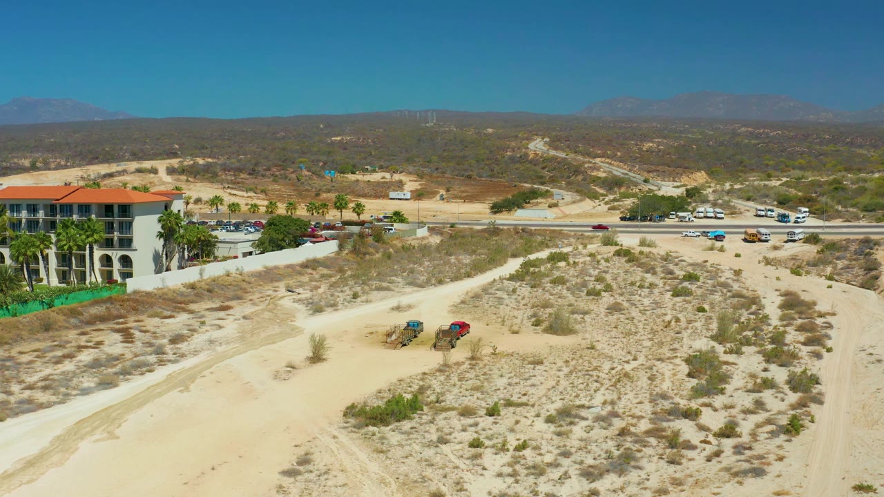 Aerial frame near Dreams Los Cabos with the red-roofed building shifted farther into view, cars on the paved road, sandy foreground tracks, low desert vegetation, mountains, and no visible people.