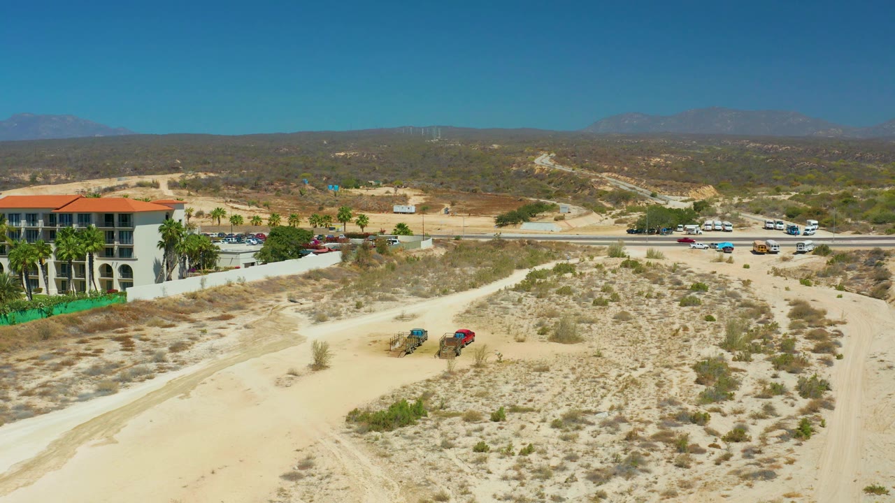 Wide aerial view near Dreams Los Cabos showing the red-roofed building on the left edge, road traffic through the middle, sandy open ground in front, distant brown hills, and no visible people.