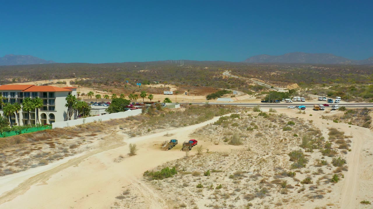Aerial view near Dreams Los Cabos with a sandy foreground, scattered desert plants, a palm-lined red-roofed building at left, vehicles along the road, hazy mountains, and no visible people.