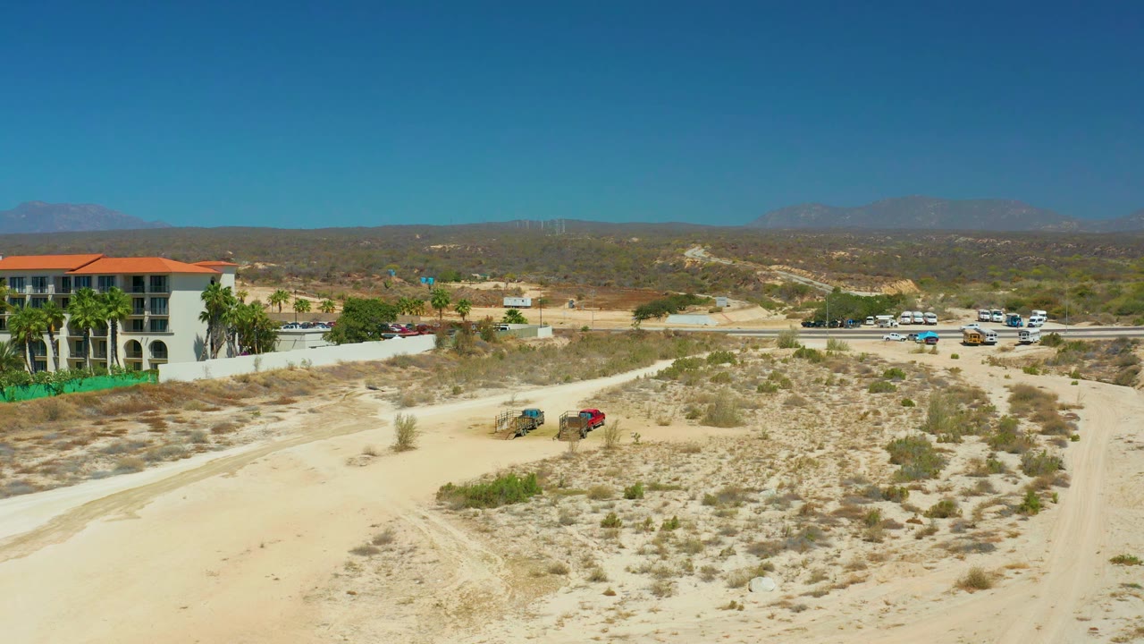 One second into the clip, the two vehicles sit farther along the dirt road through sparse desert vegetation at Dreams Los Cabos, with the red-roofed building at left and no visible people.