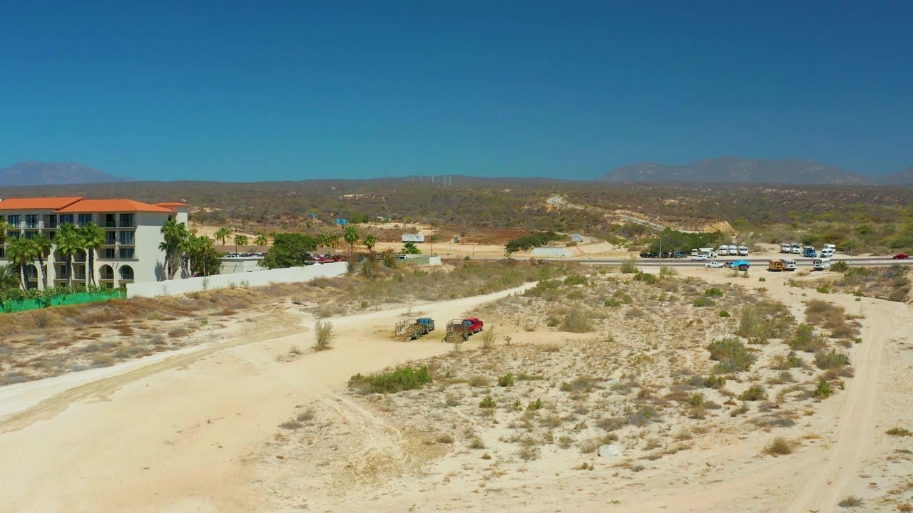 The vehicles remain on the pale dirt track below a wide blue sky, while the Dreams Los Cabos resort-edge building, parked cars, dry brush, and distant mountains stay visible with no people in frame.