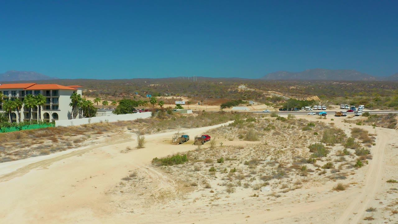 Midway through the clip, the two vehicles continue across the desert road, with low scrub in the foreground, the red-roofed white building at the left edge, parked cars beyond, and no visible people.