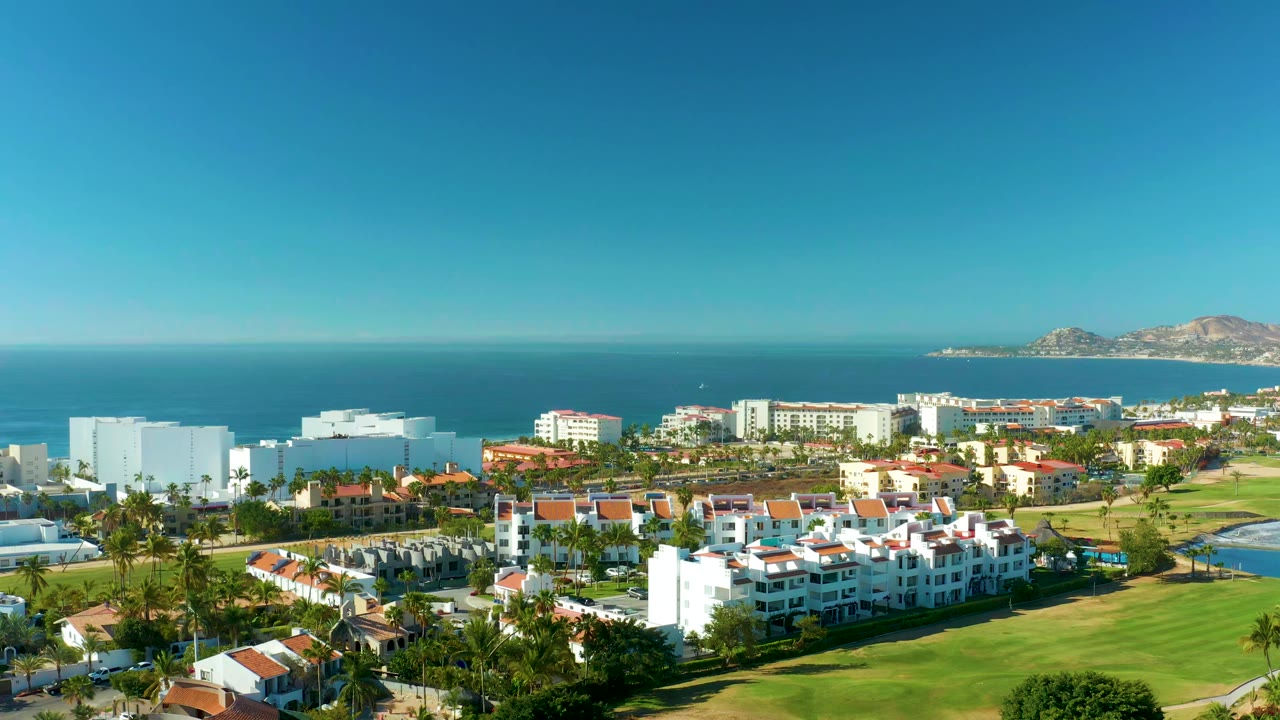 Aerial view one second into the San Jose Del Cabo pan, showing white hotel and resort blocks, red-roofed buildings, green grass, ocean, and no visible people.