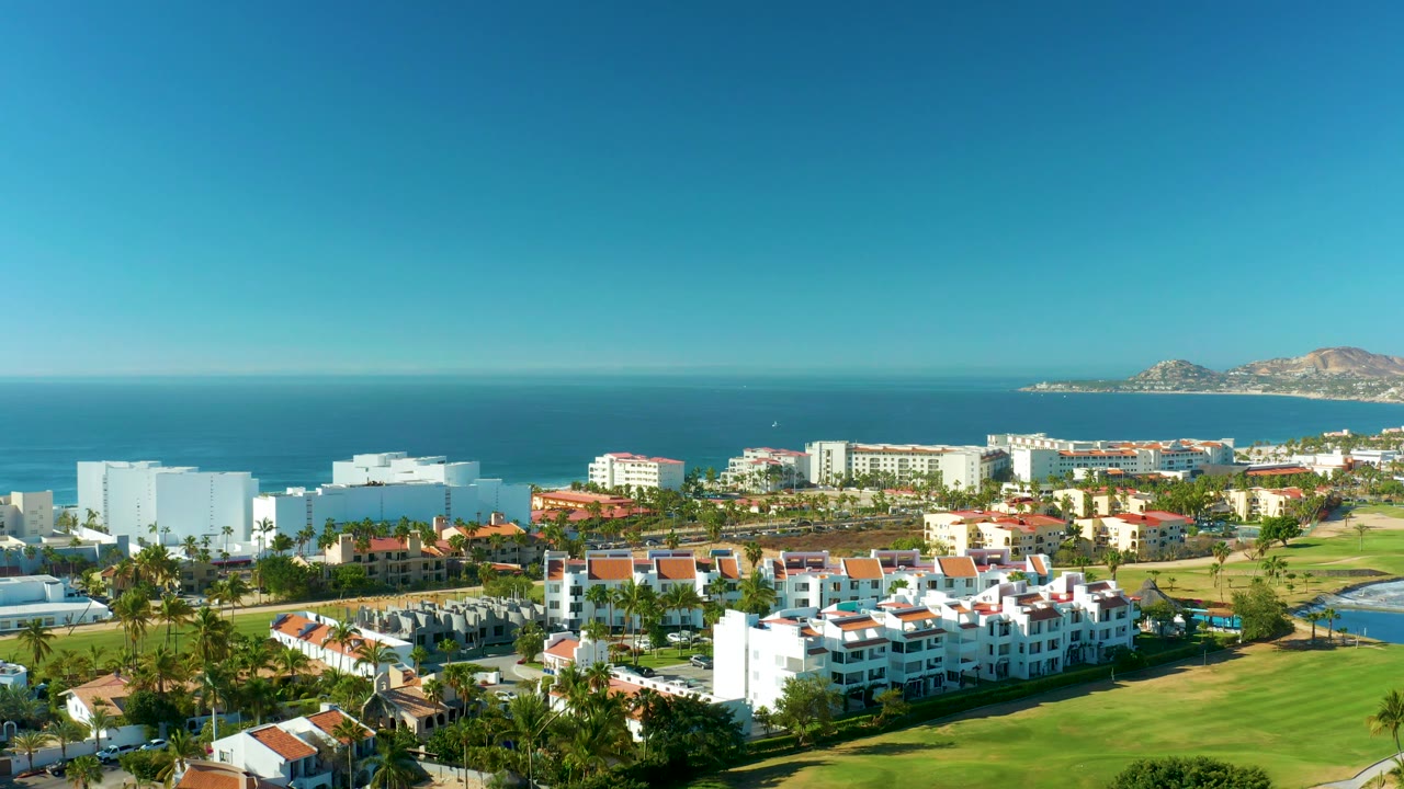 Mid-clip aerial over San Jose Del Cabo with large white buildings at left, red-roofed resort units below, green grounds in front, ocean beyond, and no visible people.