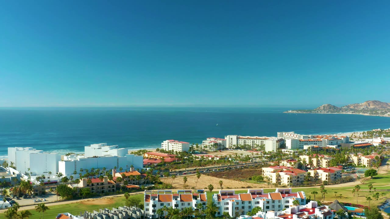 Drone view of beachfront buildings, road, palms, beach, and ocean.