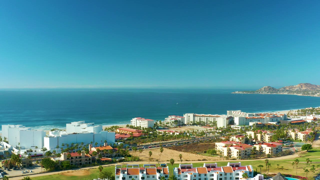 San Jose Del Cabo coastal cityscape from above, with beachfront hotels or residences, a shoreline road, surf line, open blue water, distant land at right, and no visible people.