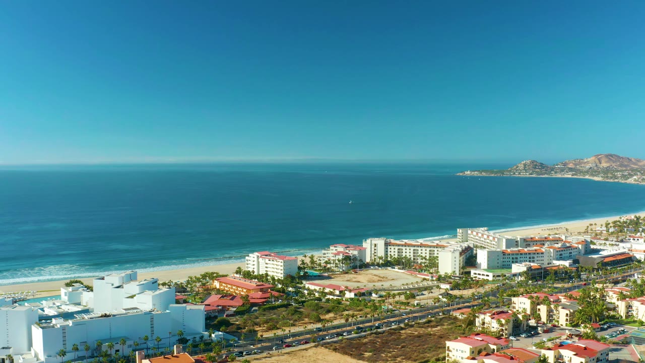 Aerial view in San Jose del Cabo with the ocean horizon high in frame, beach and surf below, white hotel-like buildings, red-roof structures, road traffic, and no visible people.