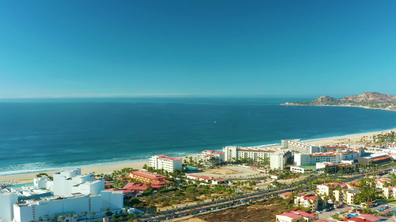 San Jose del Cabo coastal frame showing broad blue water, a bright beach strip, white multi-story beachfront buildings, a parallel road, parked vehicles, and no visible people.