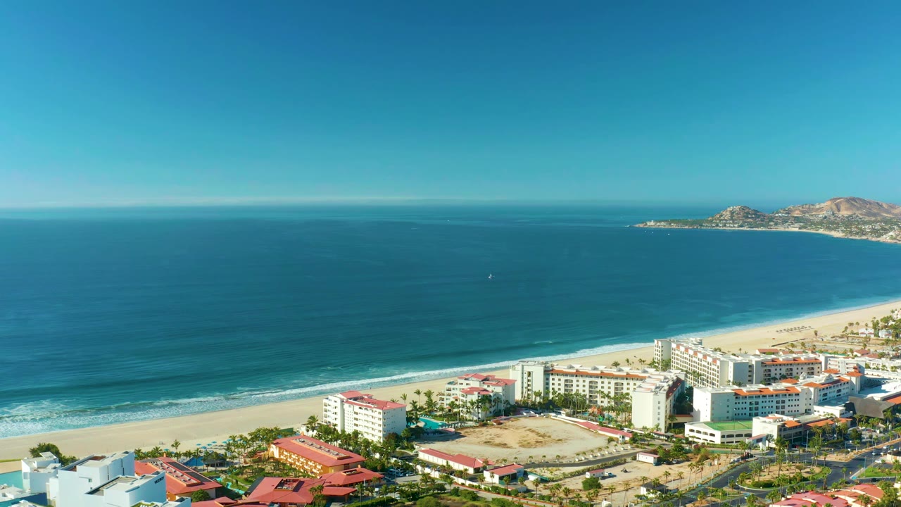 Early pan frame in San Jose Del Cabo showing more open ocean above a bright sand beach, clustered beachfront buildings, red roofs, and no visible people.