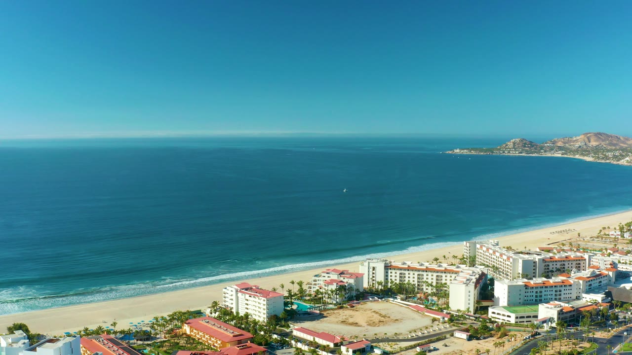 Mid-clip frame over San Jose Del Cabo with gentle waves approaching the sand, beachfront hotels or apartments along the lower edge, rocky hills in the distance, and no visible people.