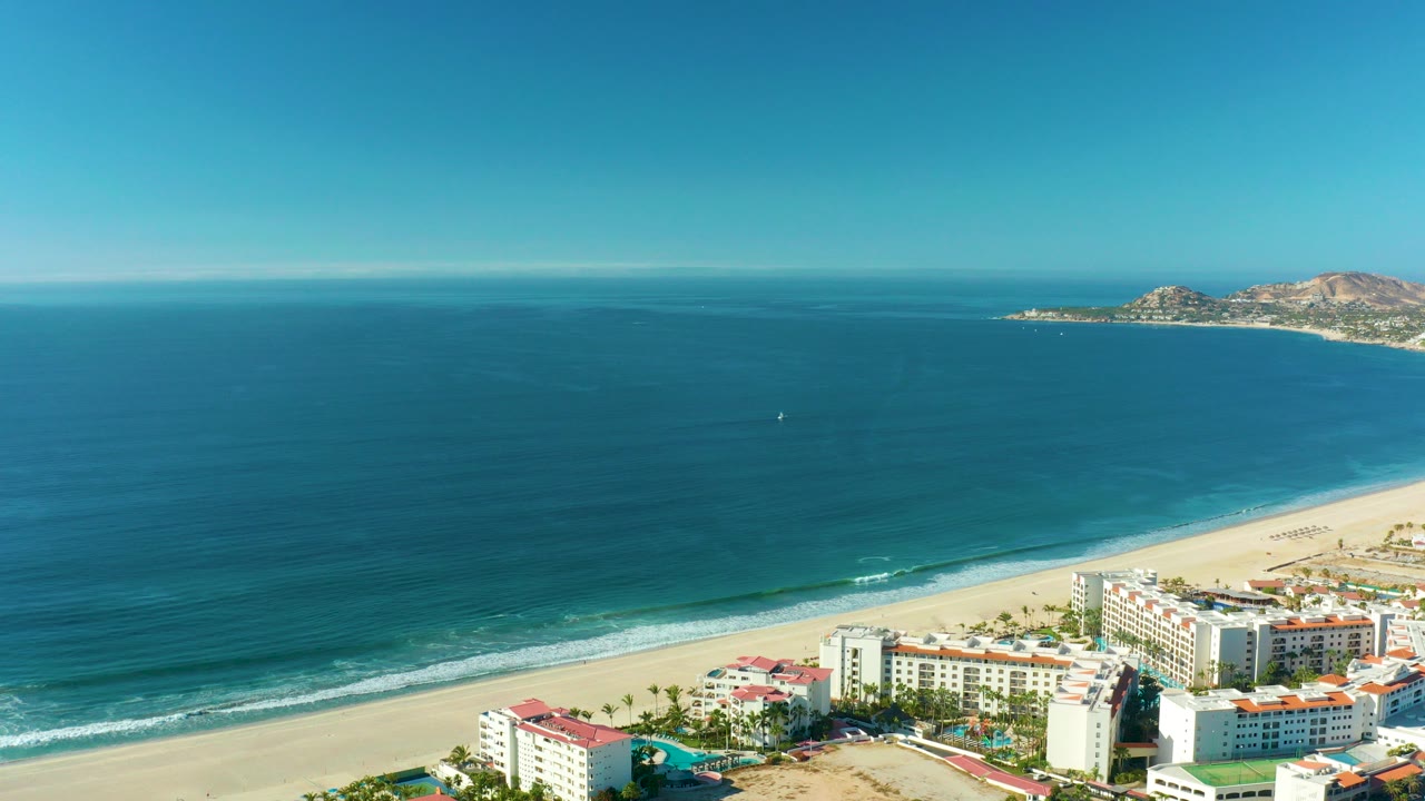 Aerial frame over San Jose del Cabo showing resort buildings at the lower edge, pale sand, calm blue ocean, distant rocky shoreline, clear sky, and no visible people.