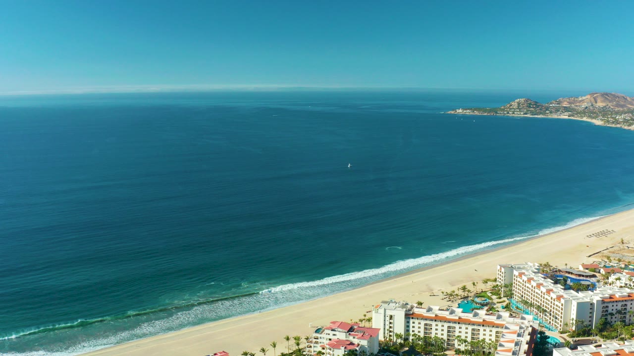 A wide aerial frame over San Jose del Cabo shows deep blue ocean, a sandy beach with white surf, coastal buildings at the lower right, a distant rocky coast, and no visible people.