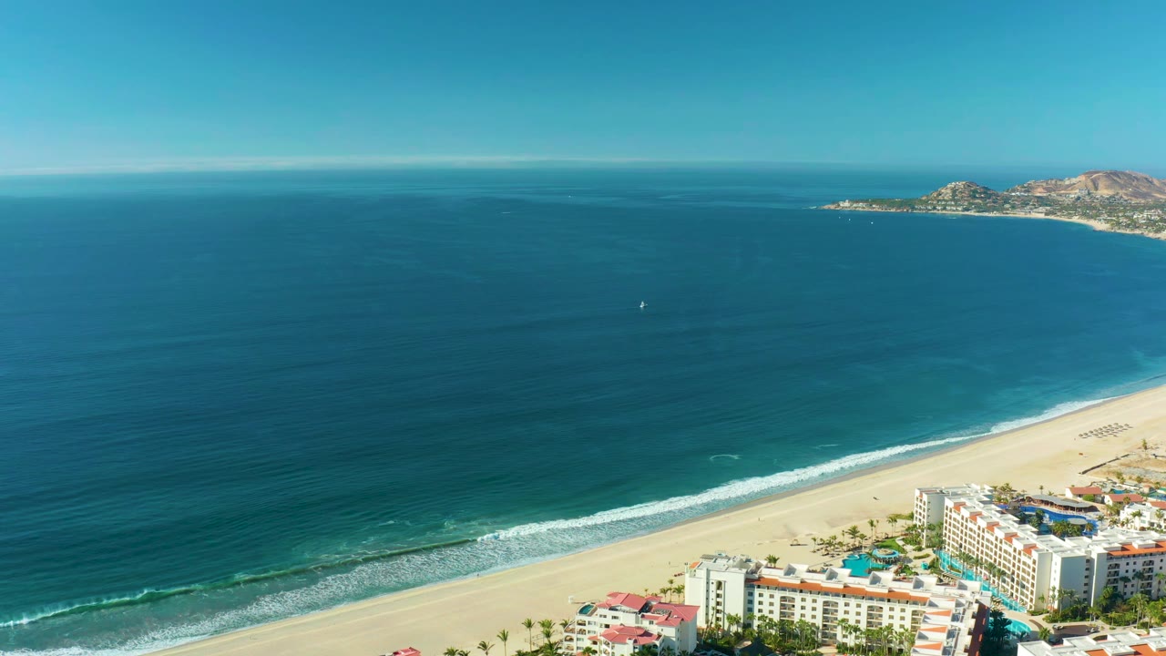 At one second, the San Jose del Cabo shoreline sits lower in frame, with turquoise water near the sand, white foam rolling in, coastal buildings at the bottom right, and no visible people.