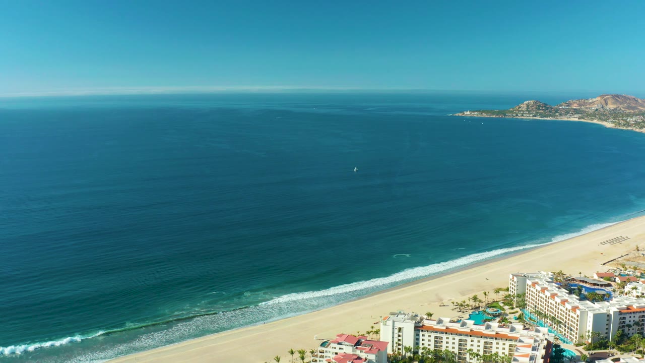 At two seconds, the aerial view keeps the horizon high, with dark blue ocean filling most of the frame, small surf bands on the San Jose del Cabo beach, buildings at the lower right, and no visible people.