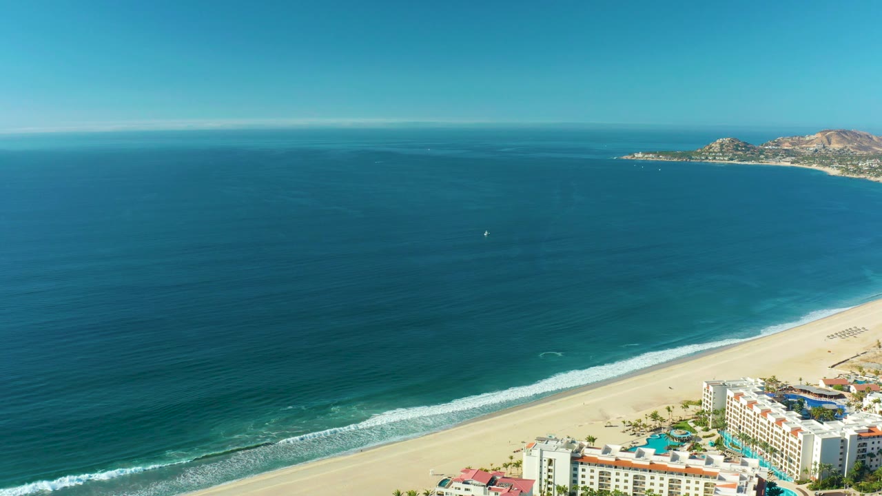 Elevated beach view with white surf, blue ocean, and a distant rocky coastline.