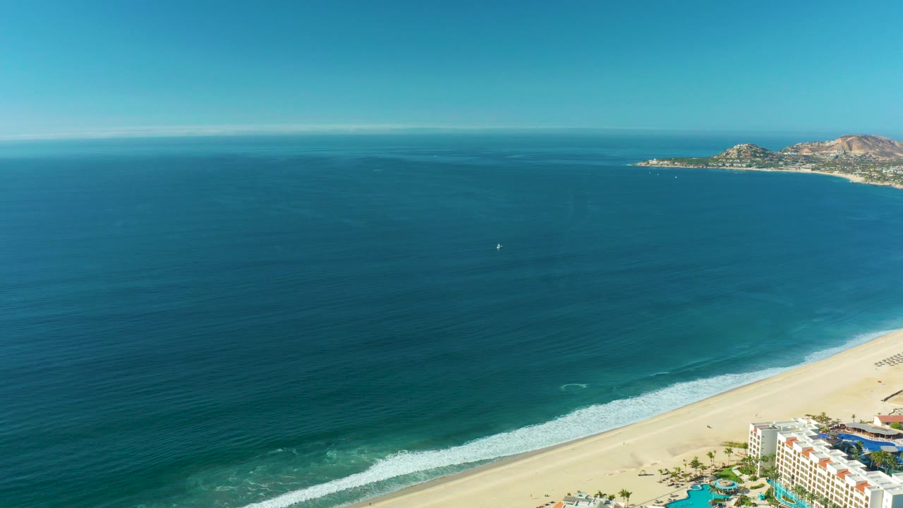 Aerial view of blue ocean waves reaching a sandy beach with distant rocky coast.
