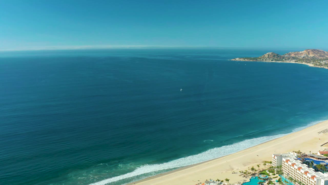 San Jose Del Cabo shoreline seen from above, with the surf line stretching diagonally across the sand and the distant coastline near the horizon, with no visible people.