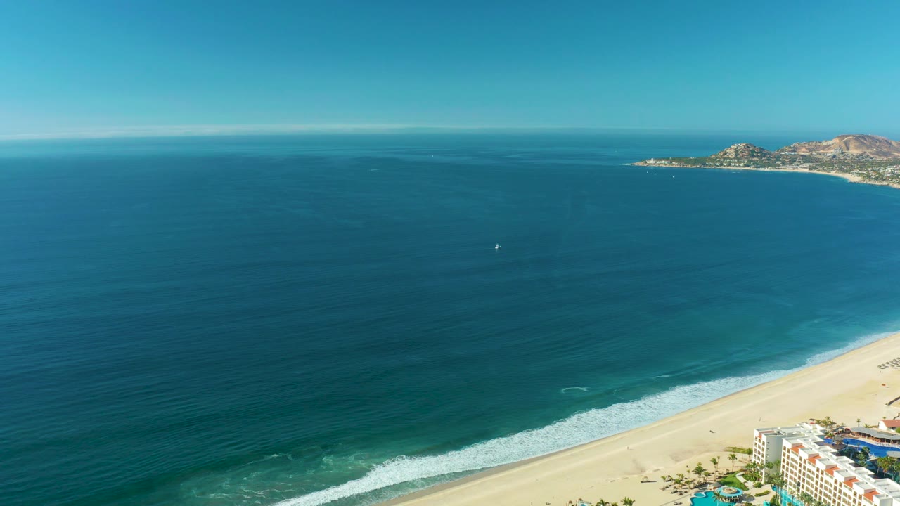 Aerial frame over San Jose Del Cabo water where deep blue sea fills the foreground and a pale beach curves along the lower right, with no visible people.