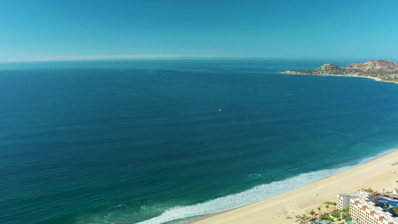 San Jose Del Cabo beach and water viewed from high above, with a thin wave band moving toward the sand and the rocky coastline set along the upper right, with no visible people.
