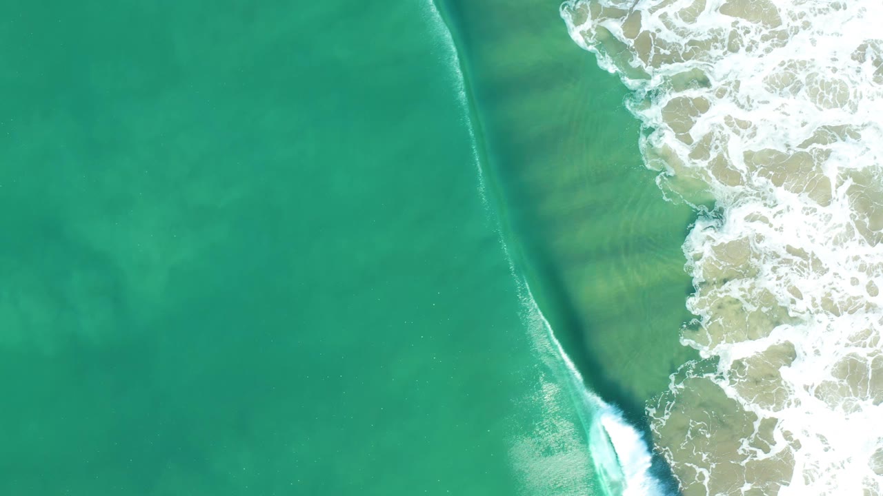 Aerial overhead frame of green-blue ocean water with foamy white waves pressing in from the right side in San Jose Del Cabo, Mexico, with no visible people.