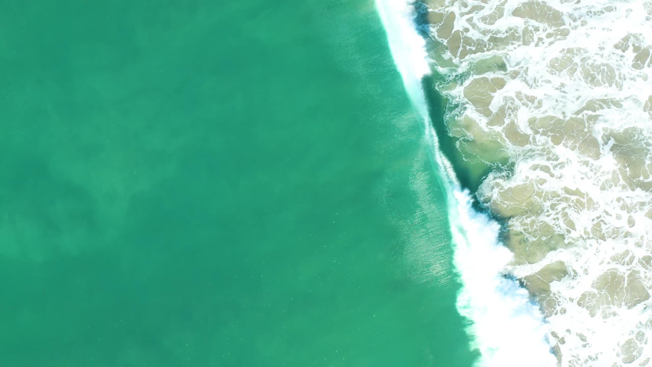 Turquoise sea fills most of the overhead frame while a bright vertical band of breaking surf runs along the right in San Jose Del Cabo, Mexico, with no visible people.