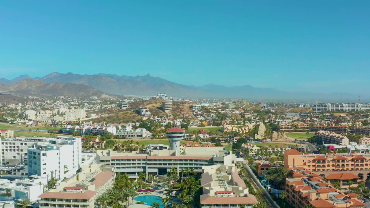 Aerial view over San Jose Del Cabo showing a white building cluster, red-roofed structures, green trees, a road corridor, and hazy mountains, with no visible people.