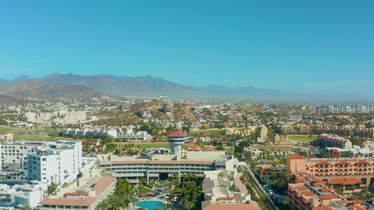 Wide San Jose Del Cabo cityscape with foreground hotel-like buildings, terracotta roofs, a small pool, scattered greenery, and a mountain range across the horizon, with no visible people.