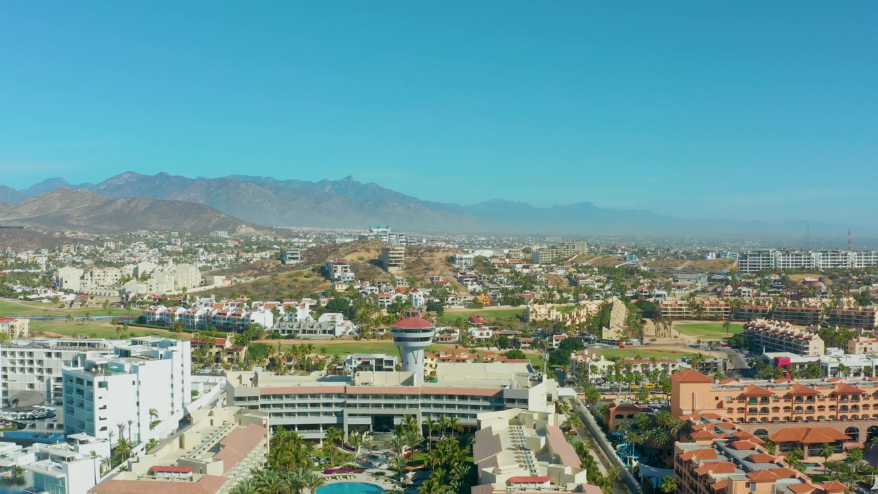 High daytime view of San Jose Del Cabo with low urban buildings, palms, white walls, red tile roofs, roads, and brown mountains beneath a cloudless sky, with no visible people.